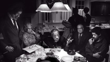 James Baldwin seated and leaning on left elbow at a table surrounded by people listening to him 